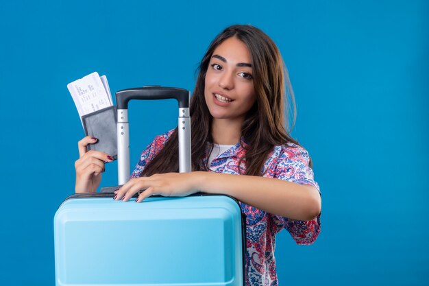 turista mulher segurando uma mala de viagem e passaporte com bilhetes com um sorriso no rosto feliz e positivo