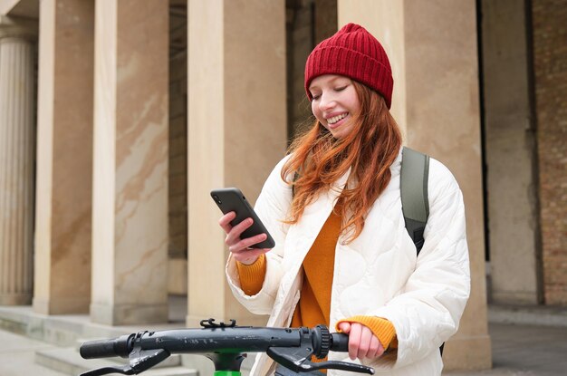 Turista ruiva com mochila usa celular para alugar escooter nas ruas da cidade europeia