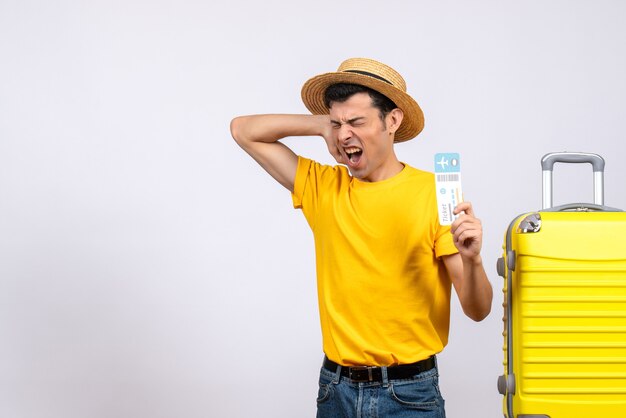 Turista zangado de frente com camiseta amarela em pé perto da mala amarela segurando o ingresso