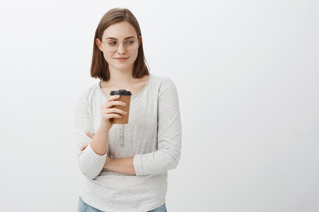 Uau, bunda linda. Retrato de uma mulher naugty, bonita e entusiasmada, com cabelo castanho e óculos, sorrindo e olhando para o canto inferior direito, bebendo café, perto de um café posando sobre uma parede cinza