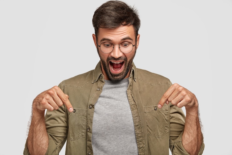 Foto grátis uau, isso é incrível! homem jovem chocado e espantado olha com surpresa para baixo, aponta com os dois dedos indicadores, vestido casualmente, abre a boca amplamente em espanto, isolado sobre a parede branca