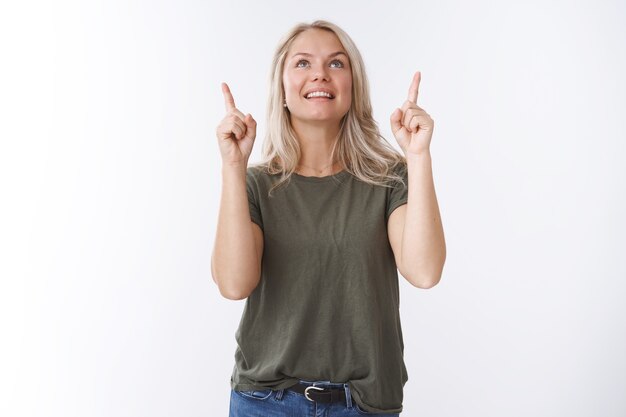 Uau que divertido. Retrato de uma jovem mulher bonita fascinada e alegre com cabelo loiro olhando apontando para cima sorrindo de emoção e alegria amorosa olhando para o céu sobre fundo branco