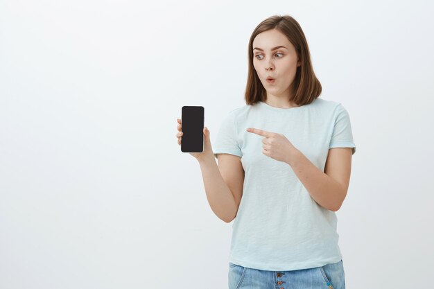 Uau, veja o que eu tenho. Menina bonita e entusiasmada impressionada com cabelo castanho curto dobrando os lábios em espanto apontando para a tela do dispositivo enquanto segura e olha para um smartphone legal sobre uma parede branca