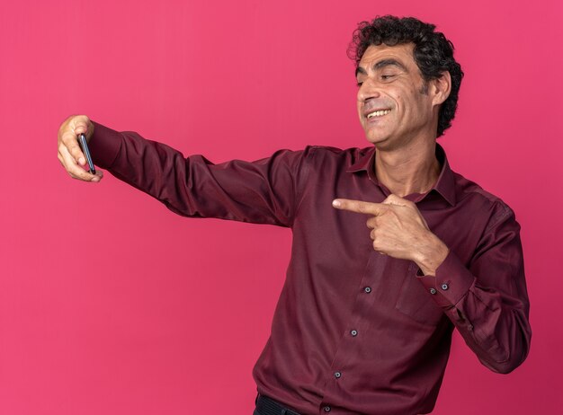 Último homem de camisa roxa fazendo selfie usando smartphone feliz e positivo sorrindo alegremente em pé sobre rosa