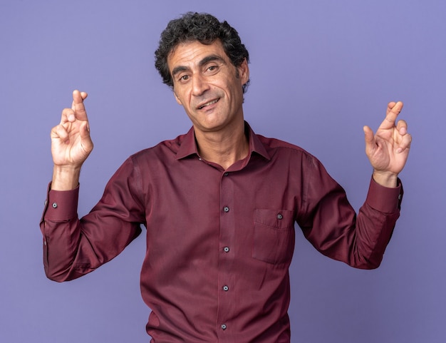 Foto grátis Último homem de camisa roxa olhando para a câmera sorrindo e fazendo um desejo cruzando os dedos em pé sobre um fundo azul