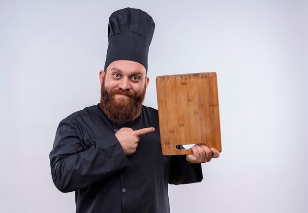 Um alegre chef barbudo de uniforme preto apontando para a placa de madeira da cozinha em uma parede branca