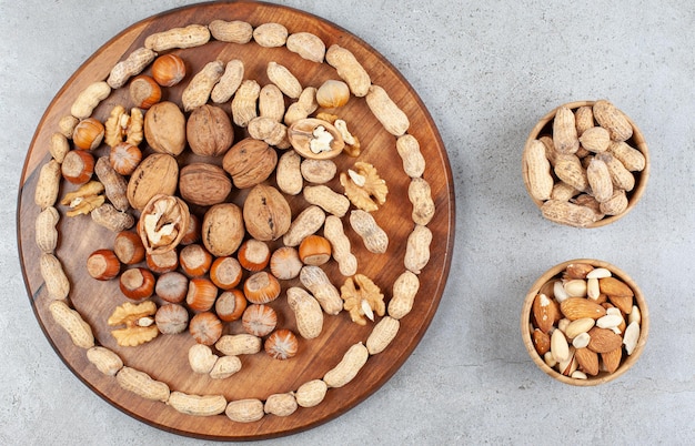 Um arranjo de vários tipos de nozes em uma placa de madeira com tigelas de amendoim, amêndoas e pistache na superfície de mármore.