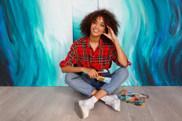 Um artista de sorriso novo da mulher negra no estúdio que guardara uma escova. Aluno inspirado sentado sobre suas obras de arte.
