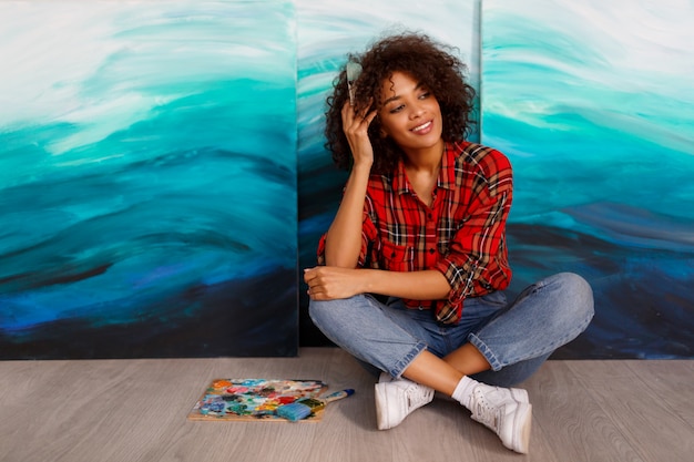 Um artista de sorriso novo da mulher negra no estúdio que guardara uma escova. Aluno inspirado sentado sobre suas obras de arte.
