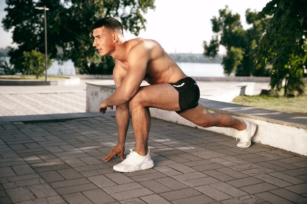 Um atleta masculino musculoso fazendo exercícios no parque. Ginástica, treino, flexibilidade de treino de fitness. Cidade de verão em dia ensolarado no campo espacial