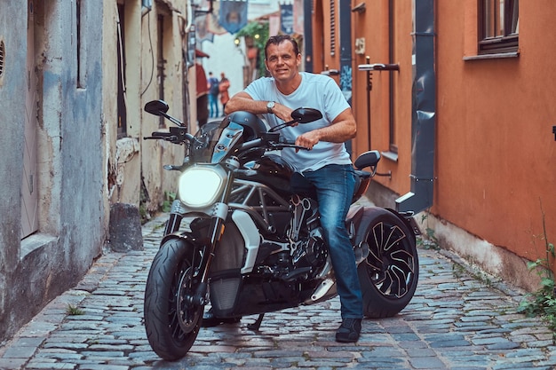 Um belo motociclista brutal vestido com uma camiseta branca e jeans, com um sorriso encantador, sentado em uma motocicleta, em uma rua estreita da velha Europa.