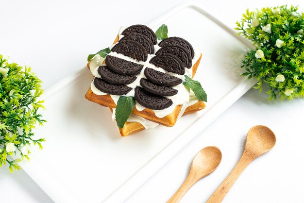 Um bolo de biscoito de vista frontal com creme e biscoitos de chocolate deliciosa sobremesa dentro da mesa branca junto com plantas e colheres de pau