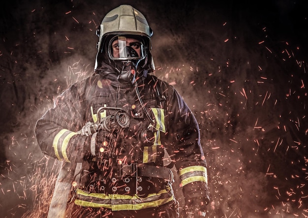 Um bombeiro profissional vestido de uniforme e uma máscara de oxigênio em pé em faíscas de fogo e fumaça sobre um fundo escuro.
