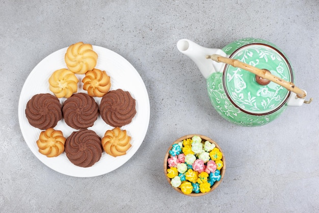 Um bule de chá ornamentado, uma tigela de doces e um prato de biscoitos na superfície de mármore