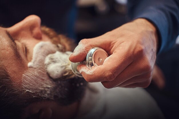 Um cabeleireiro mestre prepara o rosto para barbear e esfrega o rosto com espuma em um salão de cabeleireiro.