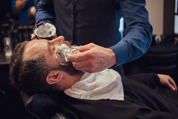 Um cabeleireiro mestre prepara o rosto para barbear e esfrega o rosto com espuma em um salão de cabeleireiro.