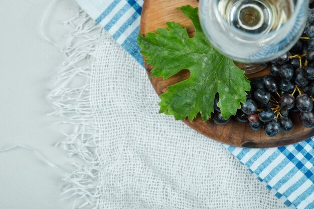 Um cacho de uvas pretas com folhas e uma taça de vinho na superfície branca