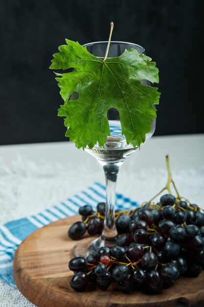 Um cacho de uvas pretas e um copo de vinho com folhas na mesa branca