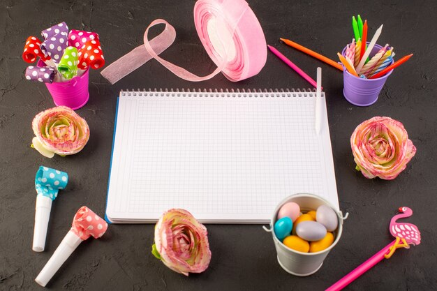 Um caderno com vista de cima e doces junto com flores, velas e lápis na mesa escura.