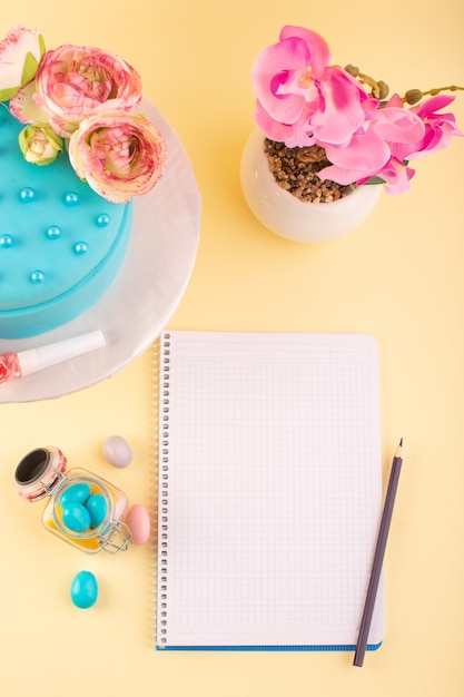 Um caderno com vista de cima e um bolo com doces e flores na mesa amarela para festa de comemoração de aniversário