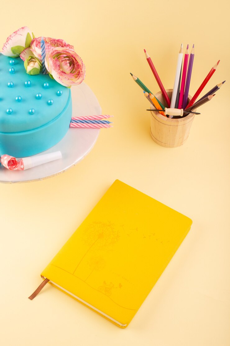 Foto grátis um caderno com vista de cima e um bolo com lápis multicoloridos na mesa amarela com a cor do bolo da celebração do aniversário