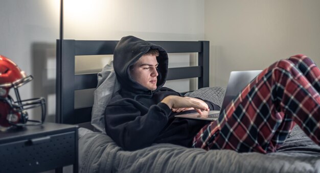 Um cara adolescente se senta em um quarto em uma cama e usa um laptop
