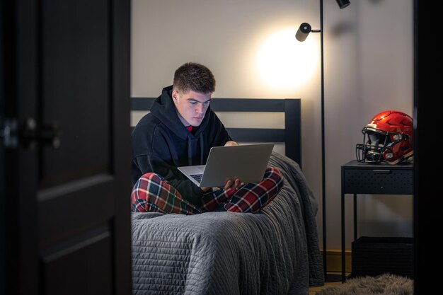 Um cara adolescente senta-se em um quarto em uma cama e usa um laptop
