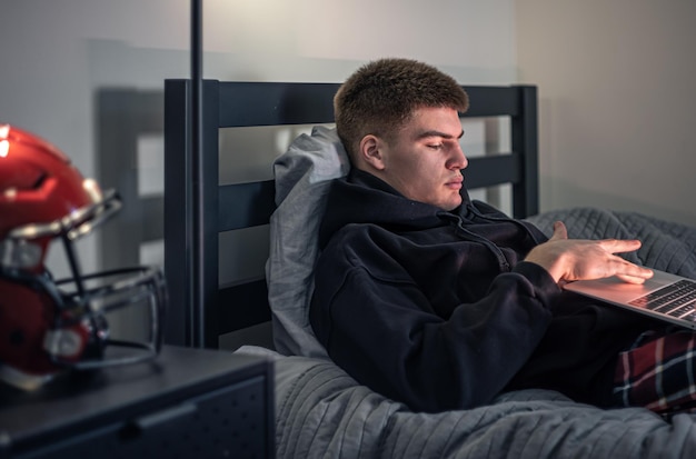 Um cara adolescente senta-se em um quarto em uma cama e usa um laptop