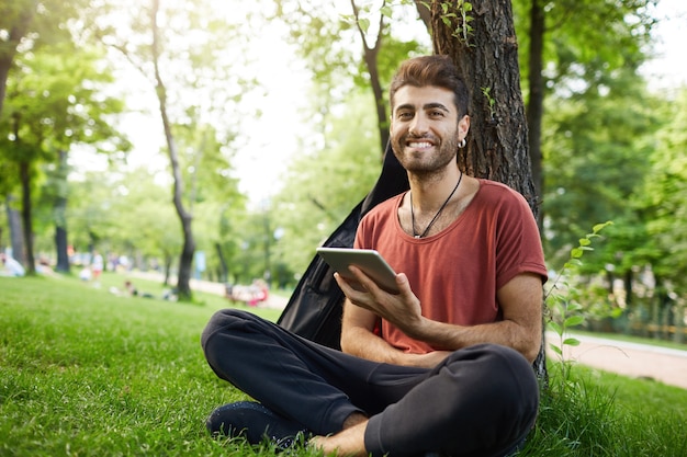 Um cara bonito sentado na grama do parque, lendo um livro digital do tablet, conectando-se ao Wi-Fi e procurando nas redes sociais