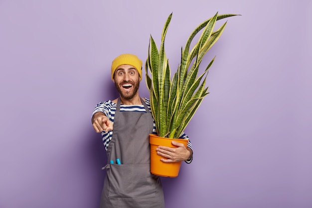 Um cara com a barba por fazer satisfeito aponta para a frente, tem um sorriso largo, mostra os dentes brancos, carrega um vaso de plantas, usa chapéu e avental amarelos