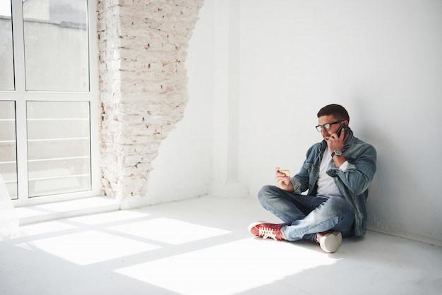 Um cara com roupas casuais está sentado em casa em um apartamento vazio e ligando para o telefone.
