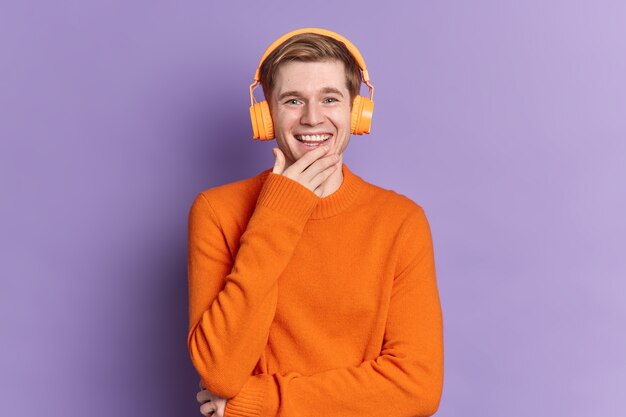 Um cara europeu bonito sorrindo e expressando emoções positivas ouvindo trilha de áudio via fones de ouvido estéreo usando jumper laranja