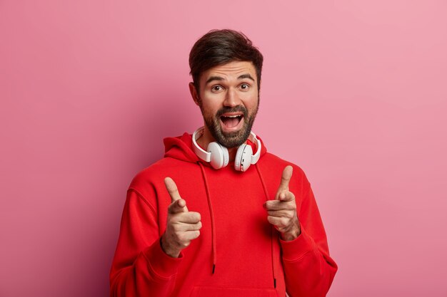 Um cara hipster barbudo positivo aponta diretamente para você, faz uma boa escolha, diz excelente, faz um gesto para você, usa um capuz vermelho e fones de ouvido modernos, posa contra uma parede rosa pastel