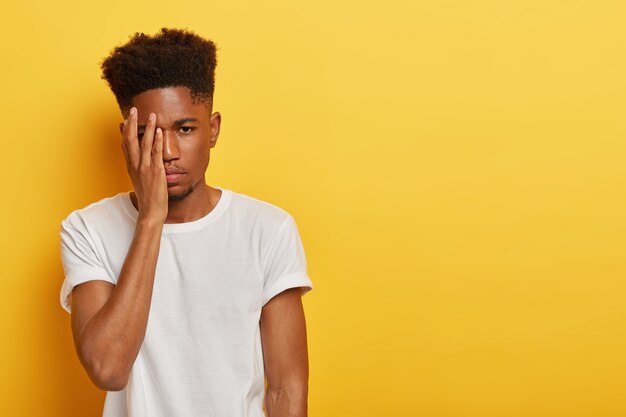 Um cara milenar carrancudo e chateado tem um penteado afro, se sente cansado do trabalho exaustivo, cobre o rosto com a palma da mão, usa uma camiseta casual branca