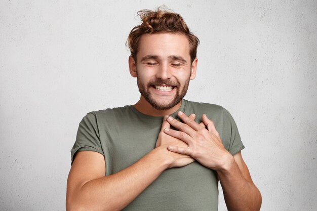 Um cara moderno feliz e bondoso sorri feliz, fecha os olhos com prazer, mantém as mãos no coração,