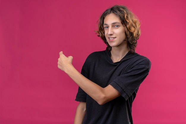 Um cara sorridente com cabelo comprido e camiseta preta aponta para as costas na parede rosa