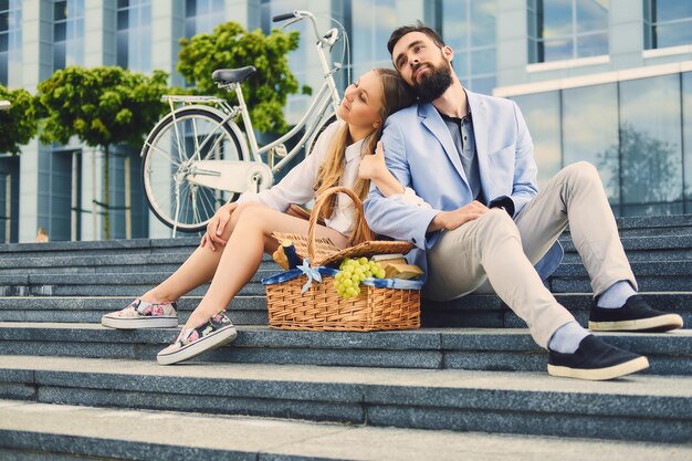 Um casal senta-se em um degrau com cesta de piquenique sobre o fundo do edifício moderno.