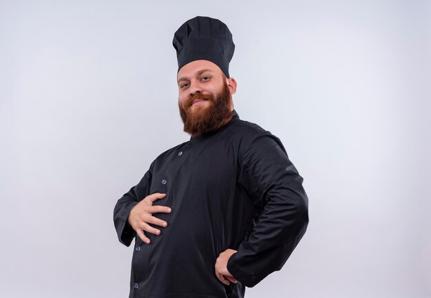 Um chef barbudo confiante em uniforme preto, segurando a mão em sua barriga enquanto segura a mão em sua cintura e olhando para uma parede branca