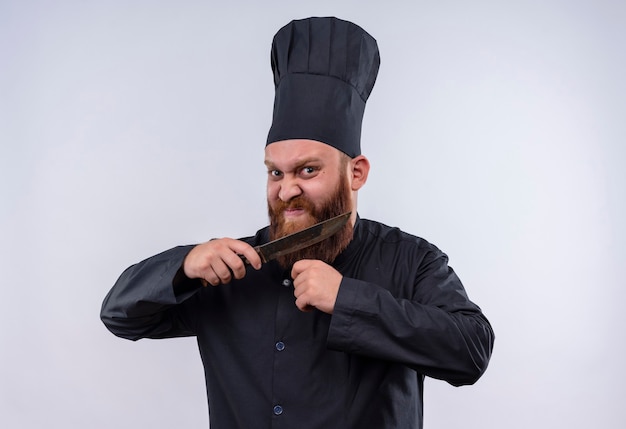 Um chef barbudo raivoso de uniforme preto segurando uma faca e tentando cortar a barba enquanto olha para a câmera em uma parede branca
