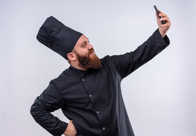 Um chef barbudo satisfeito em uniforme preto tirando uma selfie com o celular em uma parede branca