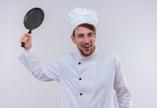 Um chef louro e alegre vestindo uniforme de fogão branco e chapéu segurando uma frigideira como um taco de beisebol em uma parede branca