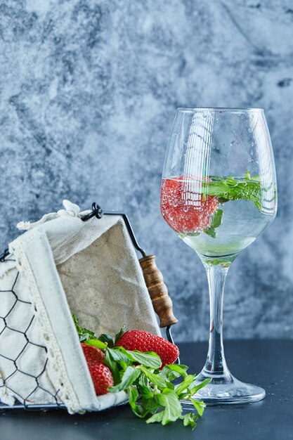 Um copo de suco com frutas inteiras dentro e uma cesta de morangos na superfície azul