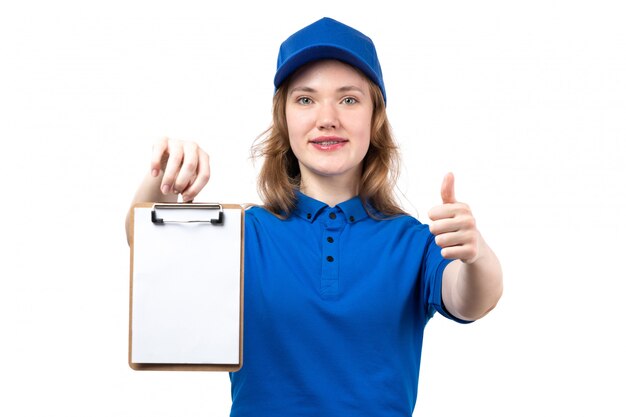 Foto grátis um correio feminino jovem vista frontal na camisa azul boné azul sorrindo mostrando como sinal e segurando o bloco de notas em branco