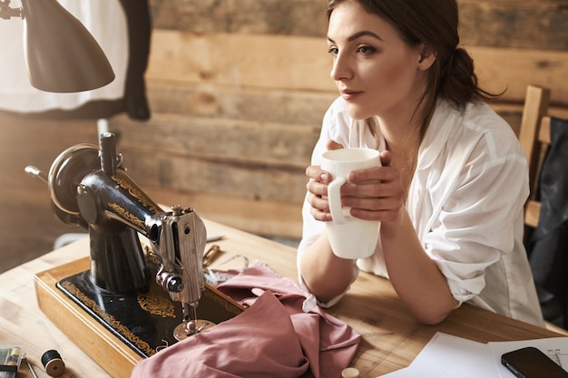 Um dia minha linha de moda se tornou famosa. um alfaiate feminino sonhador, pensando e tomando café, sentado perto da máquina de costura e do tecido, descansando enquanto cria novas roupas. o criativo prefere não se apressar