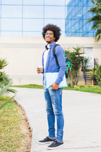 Um estudante do sexo masculino afro segurando livros e copo de café descartável em pé no campus