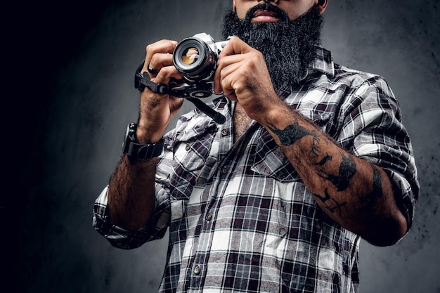 Um fotógrafo amador de hipster barbudo com tatuagens nos braços, vestido com uma camisa de lã segura uma câmera dslr compacta sobre fundo cinza.