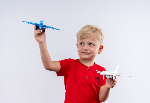 Um garotinho fofo com cabelo loiro e olhos azuis vestindo uma camiseta vermelha voando em um avião de brinquedo azul e branco enquanto olha para uma parede branca