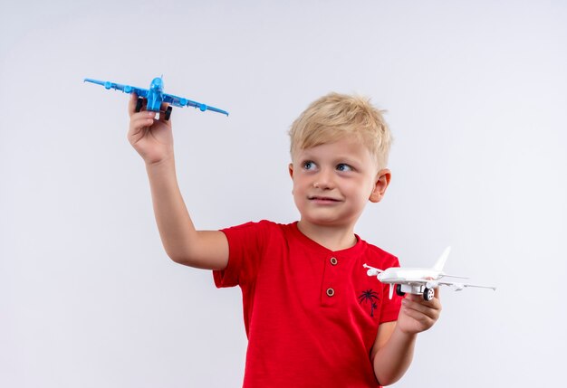 Um garotinho fofo com cabelo loiro e olhos azuis vestindo uma camiseta vermelha voando em um avião de brinquedo azul e branco enquanto olha para uma parede branca