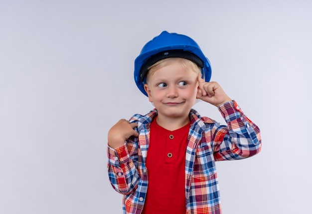 Um garotinho fofo com cabelo loiro, vestindo uma camisa xadrez com capacete azul apontando para a cabeça com o dedo indicador em uma parede branca