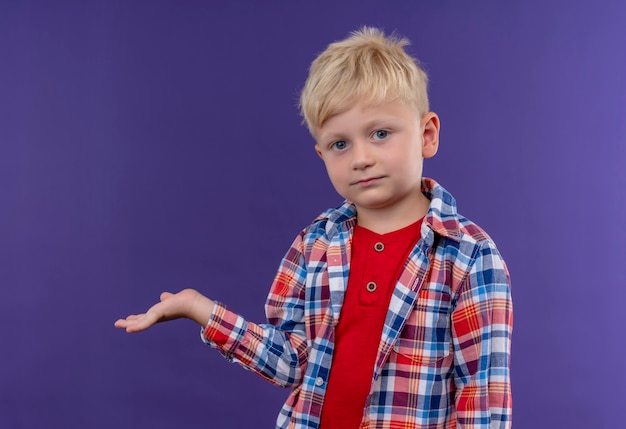 Um garotinho fofo com cabelo loiro, vestindo uma camisa xadrez, levantando a mão enquanto olha para uma parede roxa